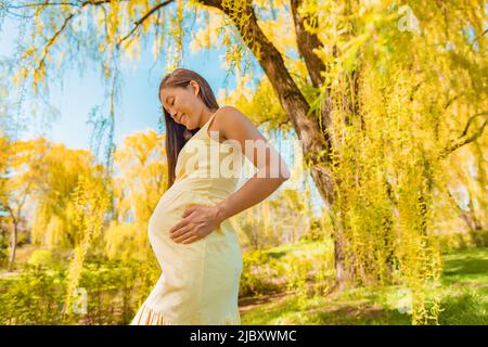 Maternity clothes pregnancy Asian pregnant woman holding baby bump belly walking outside in forest nature yellow trees wearing comfortable organic Stock Photo