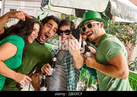 Group of friends hanging out on balcony taking a selfie and drinking beer celebrating St. Patrick's Dat. Stock Photo