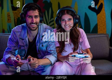 Excited friends playing video game together at home wearing microphone headsets. Stock Photo