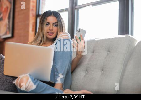 Cute Hispanic girl lounging on couch with laptop and cell phone looking into camera. Stock Photo