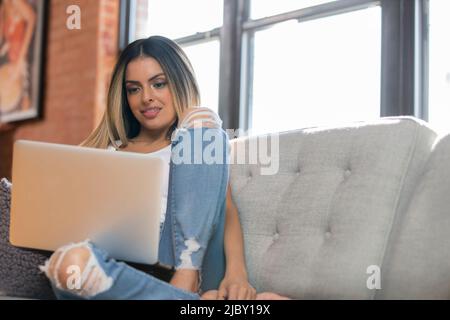 Low angle shot of Hispanic woman lounging on grey couch with laptop Stock Photo