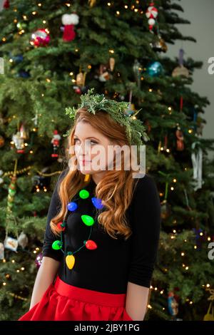 Beautiful young redhead woman standing in front of Christmas tree wearing a Christmas light necklace. Stock Photo