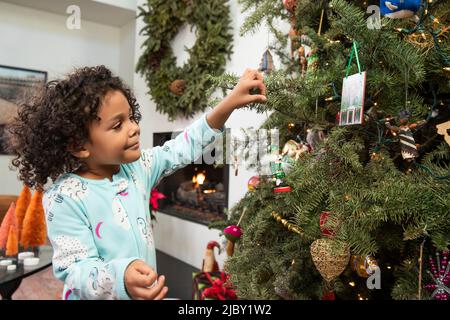 Small girl enjoying decorating and hanging ornaments on Christmas tree in living room at home. Stock Photo