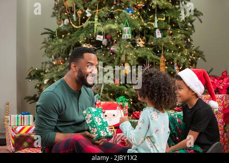 Happy father giving his young daughter a wrapped gift under Christmas tree Stock Photo