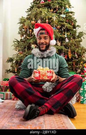 Portrait of man with Christmas gift sitting in front of tree wearing Santa hat. Stock Photo