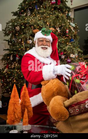 Santa Claus pulling presents out of his toy sack and placing them under Christmas tree. Black Santa Series. Stock Photo