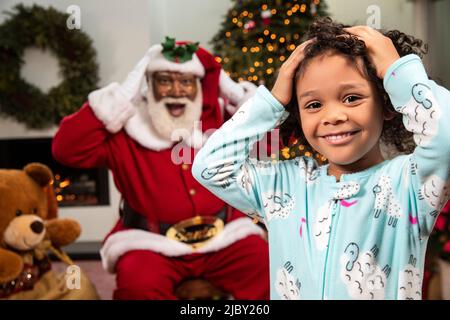 Little girl in pajamas standing by Christmas tree, with her hands on her head and Santa Claus behind her. Black Santa. Stock Photo