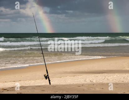 Surf caster fishing rod secured in the sand and double rainbow in stormy sky at the beach Stock Photo