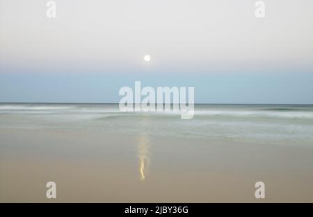 Moon rising in the evening sky over the ocean and reflecting in the sand Stock Photo