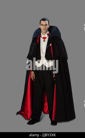 Full length portrait of a man dressed up as a vampire looking directly into camera with a spooky expression, against a gray background Stock Photo