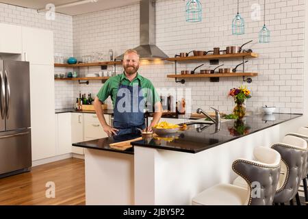 Portrait of a man with beard in domestic kitchen wearing apron and looking at camera with hands on hips. Stock Photo