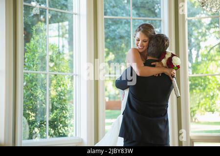 Bride and groom in an embrace, groom lifting up bride standing by large bank of windows Stock Photo