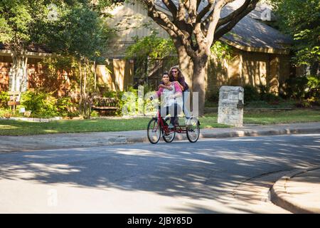 Young couple riding large tricycle through neighborhood, girl standing on back wearing heart shaped sunglasses with her arms around boyfriend Stock Photo