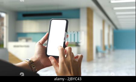 A female hands using a modern smart mobile phone over blurred hospital or clinic reception room in the background. Phone blank screen mockup. closeup Stock Photo