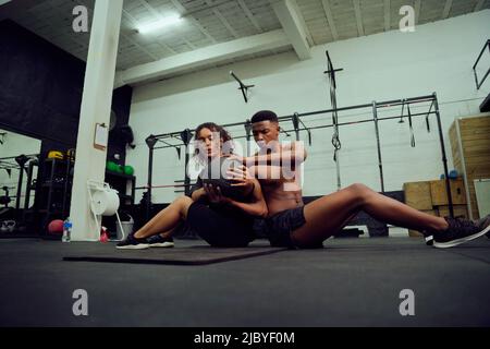 Mixed race friends doing cross fit in the gym. African American male and female sitting down on the floor and passing a medicine ball during exercise Stock Photo