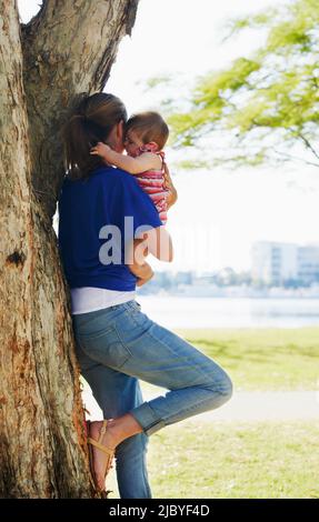 Mother leaning against tree holding baby at park Stock Photo