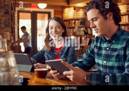 Young man and woman sitting at counter in cafe bookstore using digital tablets , woman looking at camera smiling Stock Photo