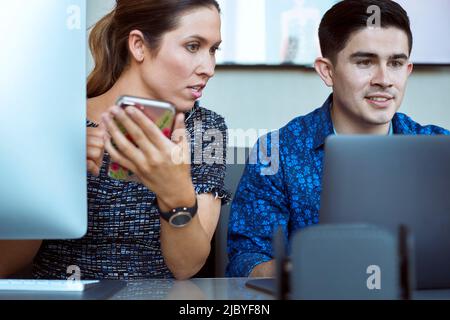 Young man and woman co-workers gathered around laptop computer looking at screen, woman checking smart phone Stock Photo