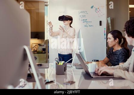 -Workers working on computer in office while woman is using Virtual Reality Headset standing in front of dry erase board with flowchart in background Stock Photo