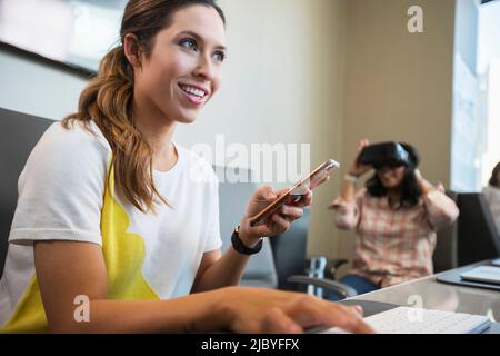 Young woman in conference room working on computer checking mobile phone, with co-worker in background wearing a Virtual reality headset Stock Photo
