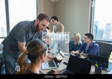 worker in office conference room providing tech support to colleague working on laptop, co-workers working in background Stock Photo