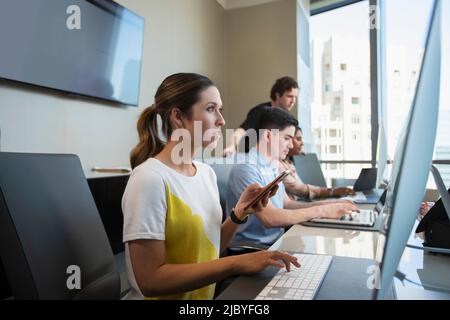 Young woman using computer in office typing on keyboard, and checking mobile phone with co-workers working in background Stock Photo