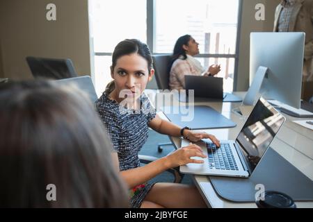 Woman taking to co-worker while working on laptop computer, other colleagues having discussion in background Stock Photo