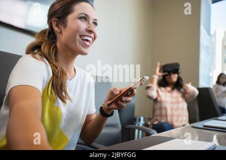 Young woman in conference room working on computer checking mobile phone, with co-worker in background wearing a Virtual reality headset Stock Photo