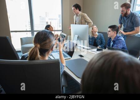 Woman taking selfie of herself and co-worker with mobile phone, while colleagues work in background of conference room Stock Photo