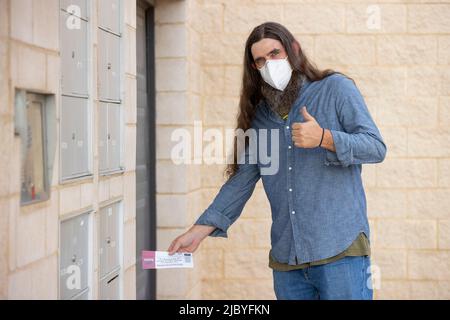 an male with long hair and full beard wearing a mask and putting official election mail in ballot in the outgoing mail slot at his apartment building Stock Photo