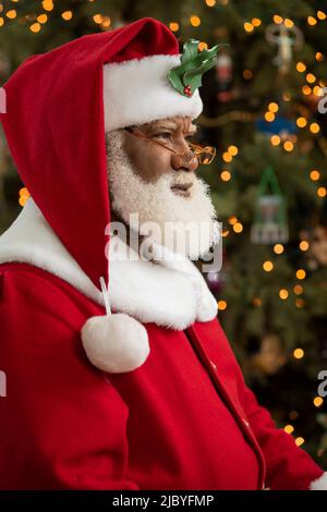 An African American man dressed as Santa Claus sitting in front of a Christmas tree turned profile, looking off camera. Stock Photo
