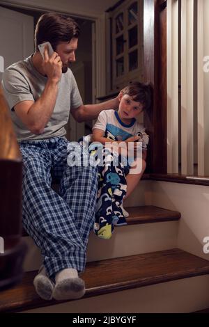 Father and son sitting at top of stairs in home getting ready for bedtime, dad calling doctor and checking on his son who is feeling ill Stock Photo