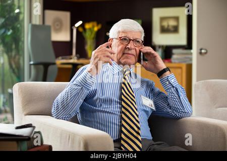 Senior Caucasian doctor practicing tele-medicine from his home office, Talking to patient on telephone Stock Photo