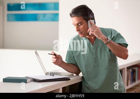 Hispanic Male doctor in his home practicing Telemedicine, talking on cell phone and taking notes Stock Photo