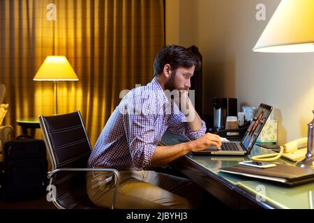 Caucasian man working late in a hotel room, feeling ill so he is having telemedicine meeting with doctor on laptop Stock Photo