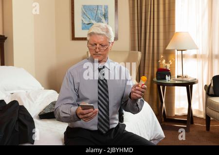 Mature Caucasian man sitting on edge of bed in a hotel room, using cell phone to check prescription medication Stock Photo