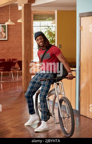 Portrait of young ethnic man wearing plaid pants and knit cap standing with bike in common area of downtown office loft Stock Photo