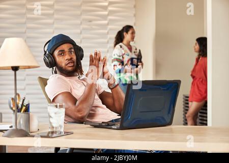 portrait young ethnic man sitting at desk with laptop computer wearing large over ear headphones, coworkers standing in background talking Stock Photo