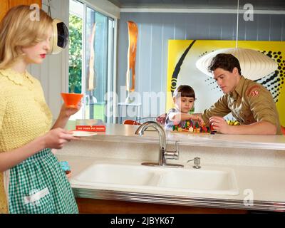 Vintage styled woman in kitchen holding a coffee mug and vintage styled man playing a board game with daughter in a mid century house. Stock Photo