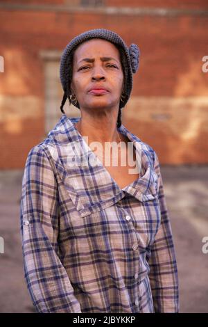 Portrait of older woman wearing knit hat standing in alley holding head up high, brick wall in background Stock Photo