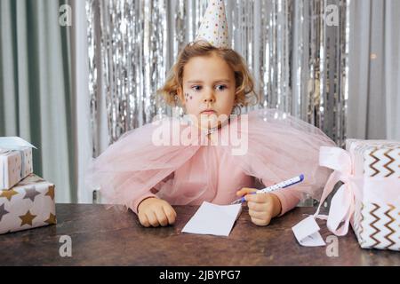 Portrait of beautiful 4-5-year-old girl with red wavy hair, dressed in party hat and pink festive outfit, who was sitting at table thinking deeply Stock Photo