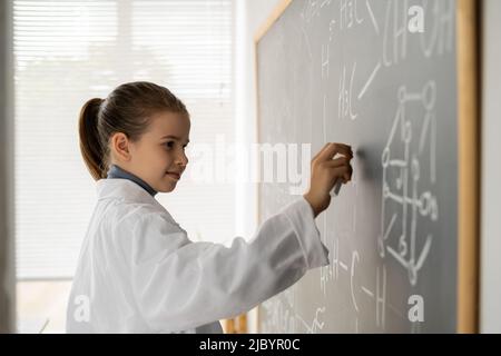 little female student in a white coat writes chemical formulas on a blackboard, science and education Stock Photo