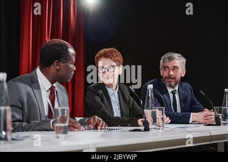 Group of business people sitting at table with microphones and talking to each other during conference Stock Photo