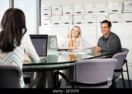 Three co-workers talk while on their computers and tablets. Stock Photo