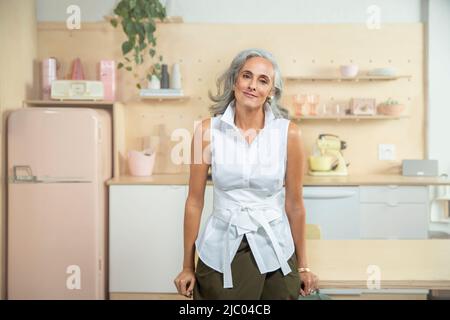 Wide horizontal portrait of a middle-aged woman with gray hair, standing in a kitchen looking into camera with sincere smile. Stock Photo