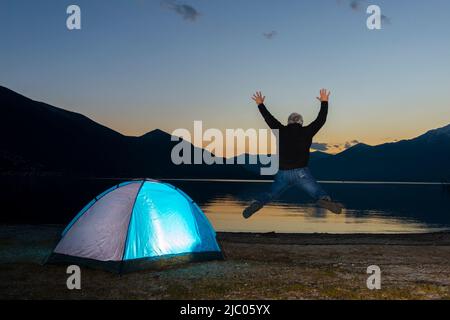 Man Jumping and an Illuminated Tent on Alpine Lake Maggiore with Mountain in Dusk in Ascona, Switzerland. Stock Photo