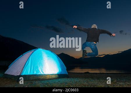 Man Jumping and an Illuminated Tent on Alpine Lake Maggiore with Mountain in Dusk in Ascona, Switzerland. Stock Photo