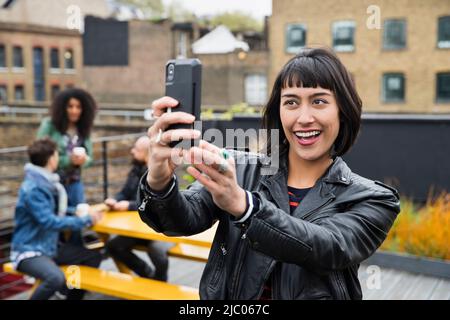 Portrait of Woman taking selfie with group of friends working on tablets at outdoor patio table in co-working space in the background Stock Photo