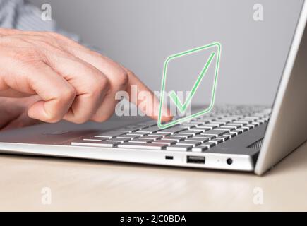 Hand at laptop keyboard with tick icon. Approval of smth, idea, project or online verification of product, service, system or data confirmation. Man forefinger pressing enter button. photo Stock Photo