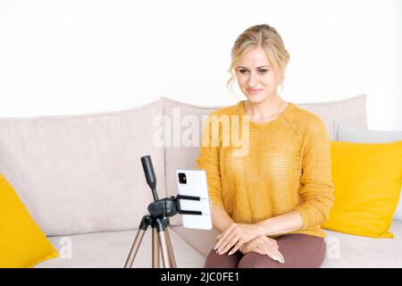 Cheerful female blogger with mobile phone tells about income to subscribers sitting on comfortable sofa in living room smiling caucasian woman gesturing with emotional face Stock Photo